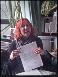 Woman sitting in office, holding a paper up to the camera and smiling broadly