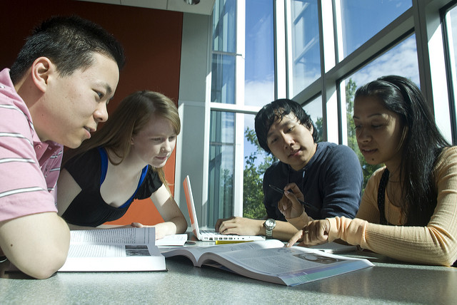 Four college students, smiling, leaning over papers on a table in a sunny room