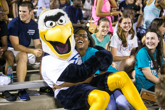 Photo of a Eagle mascot sitting on the lap of a woman in bleachers, while people around her smile