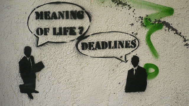 Graffiti on wall. One figure in business suit asks, "Meaning of life?" Another answers "Deadlines."