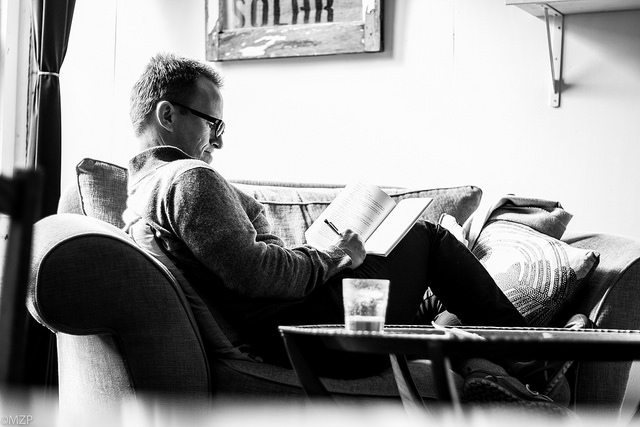 Black and white photo of a man writing in a notebook while outstretched on a couch
