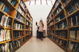 Photo of a woman holding a laptop computer standing between library stacks shelves, taken with a fishbowl lens