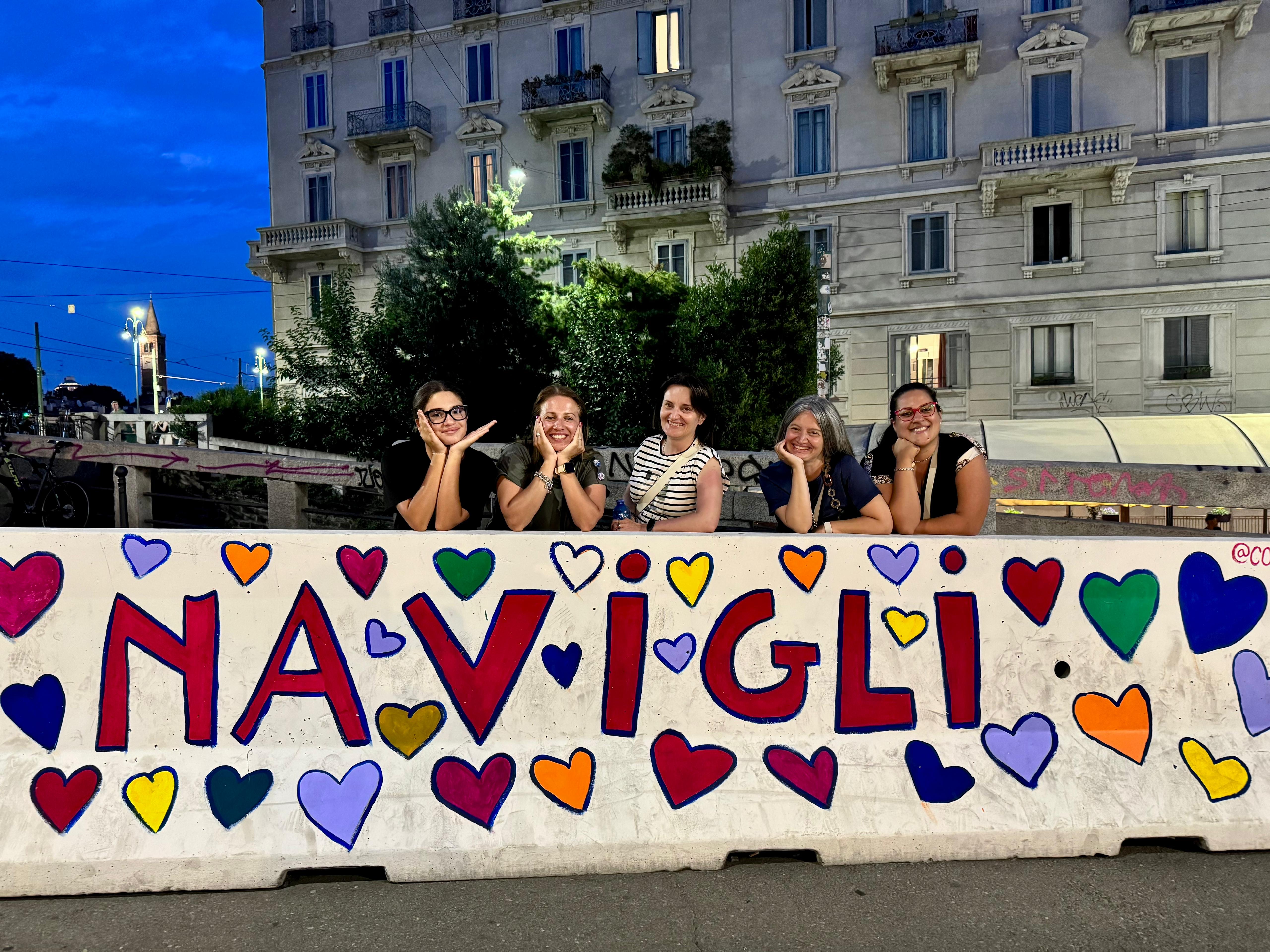 Five women pose for a photo in Milan in front of a sign that reads &ldquo;I Navigli,&rdquo; with a historic Milanese building in the background.