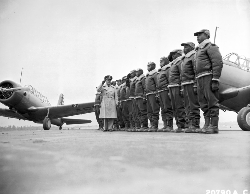 Photograph of Tuskegee Airmen standing at attention and a fighter plane in the background.