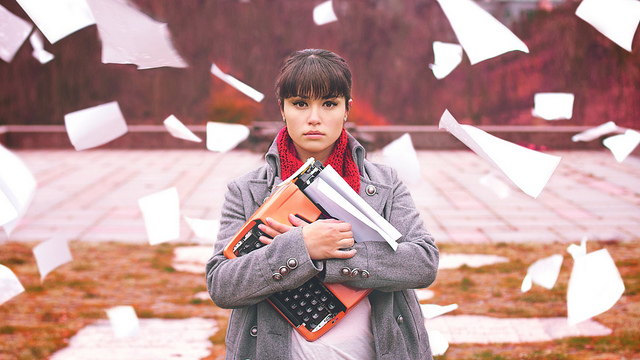 Woman holding an orange typewriter, staring at the camera, while sheets of paper blow in the wind around her