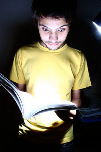 Young man in yellow shirt looking down with wide eyes at a book he's holding, which is glowing