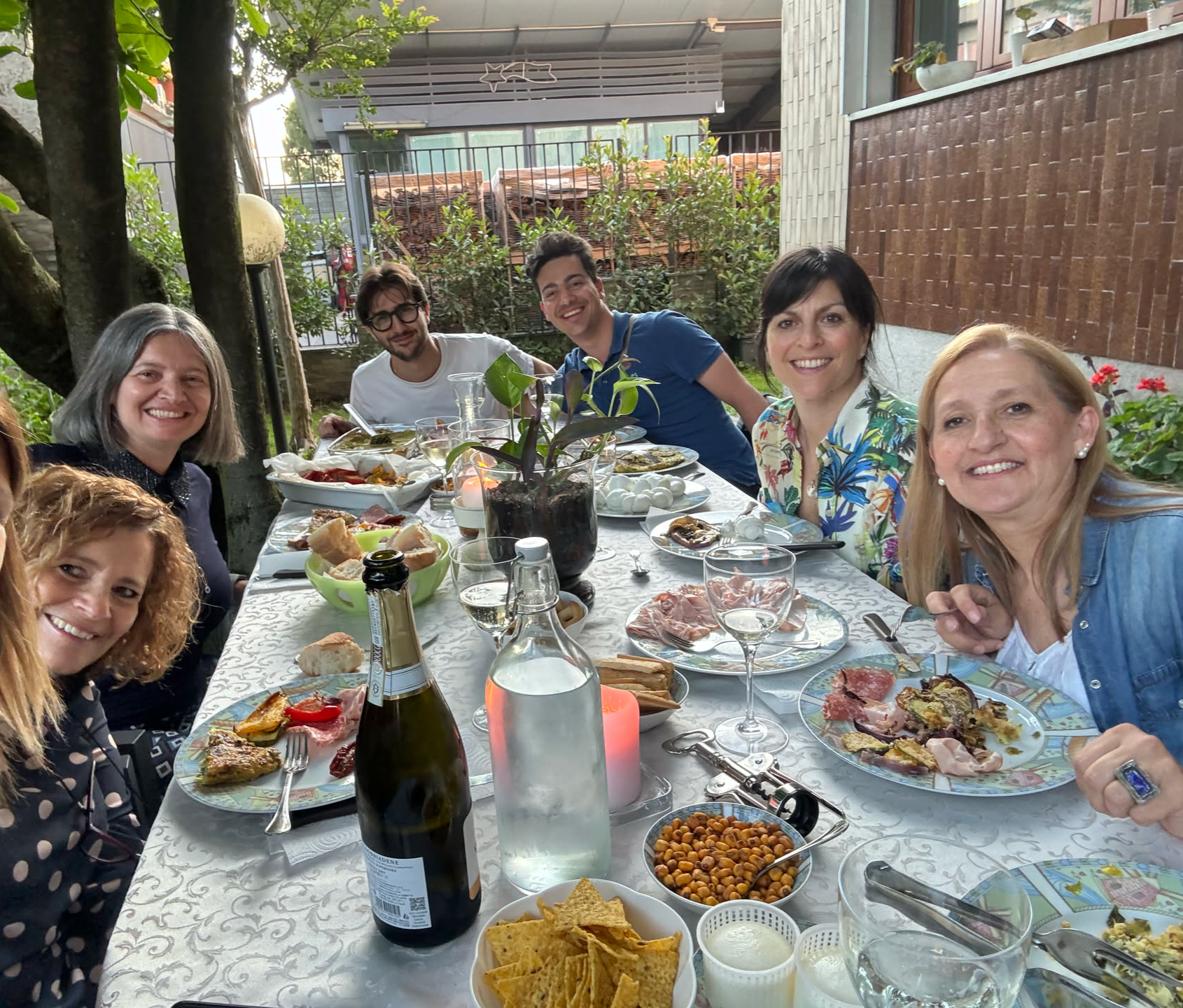 A group of seven people enjoying a meal outdoors at a table, surrounded by greenery and various dishes.