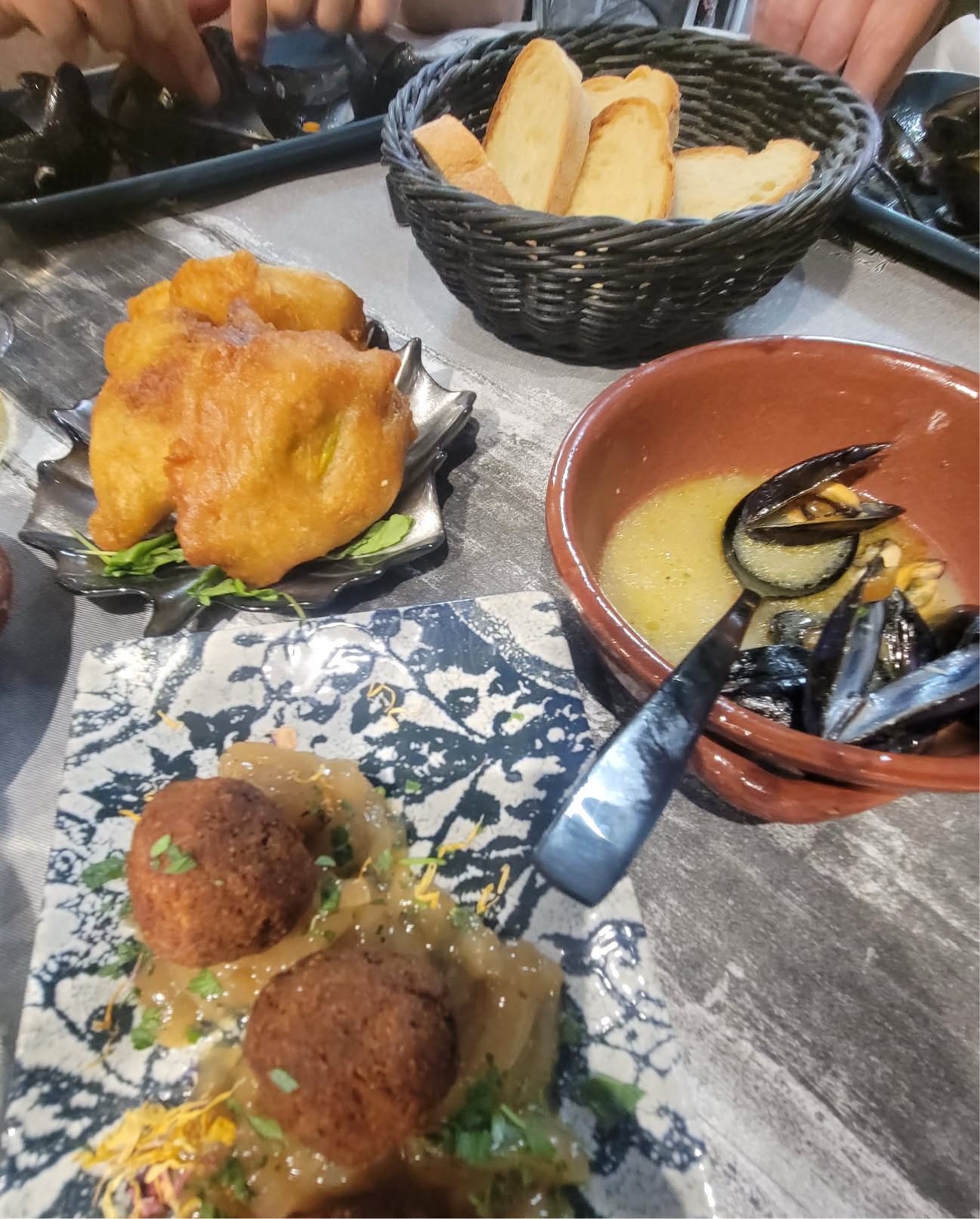 A table setting featuring fried dough, two meatballs on sauce, a bowl of mussels, and a basket of sliced bread.