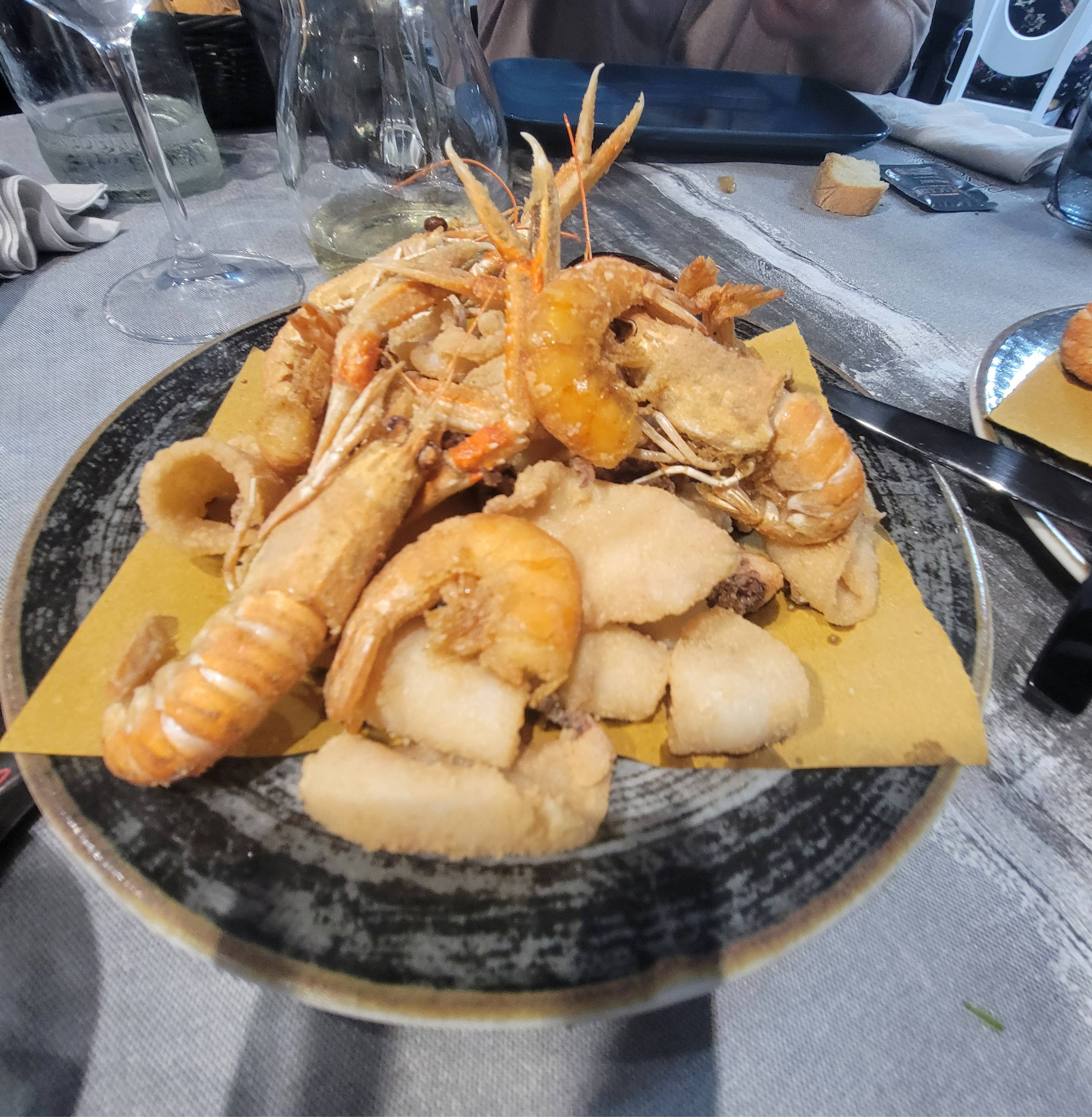 A plate of assorted fried seafood, including shrimp and small fish, served on a yellow paper on a textured dish.