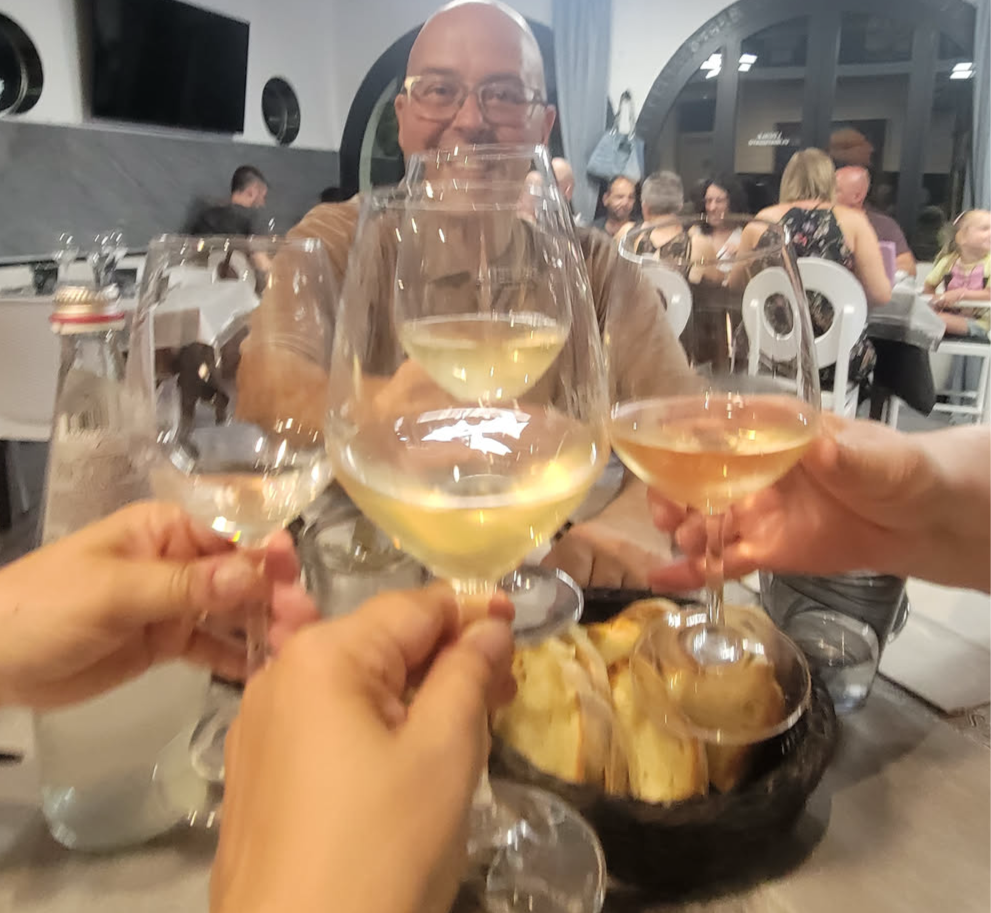 A toast with three wine glasses in the foreground, with a man smiling in the background, and a basket of bread on the table.