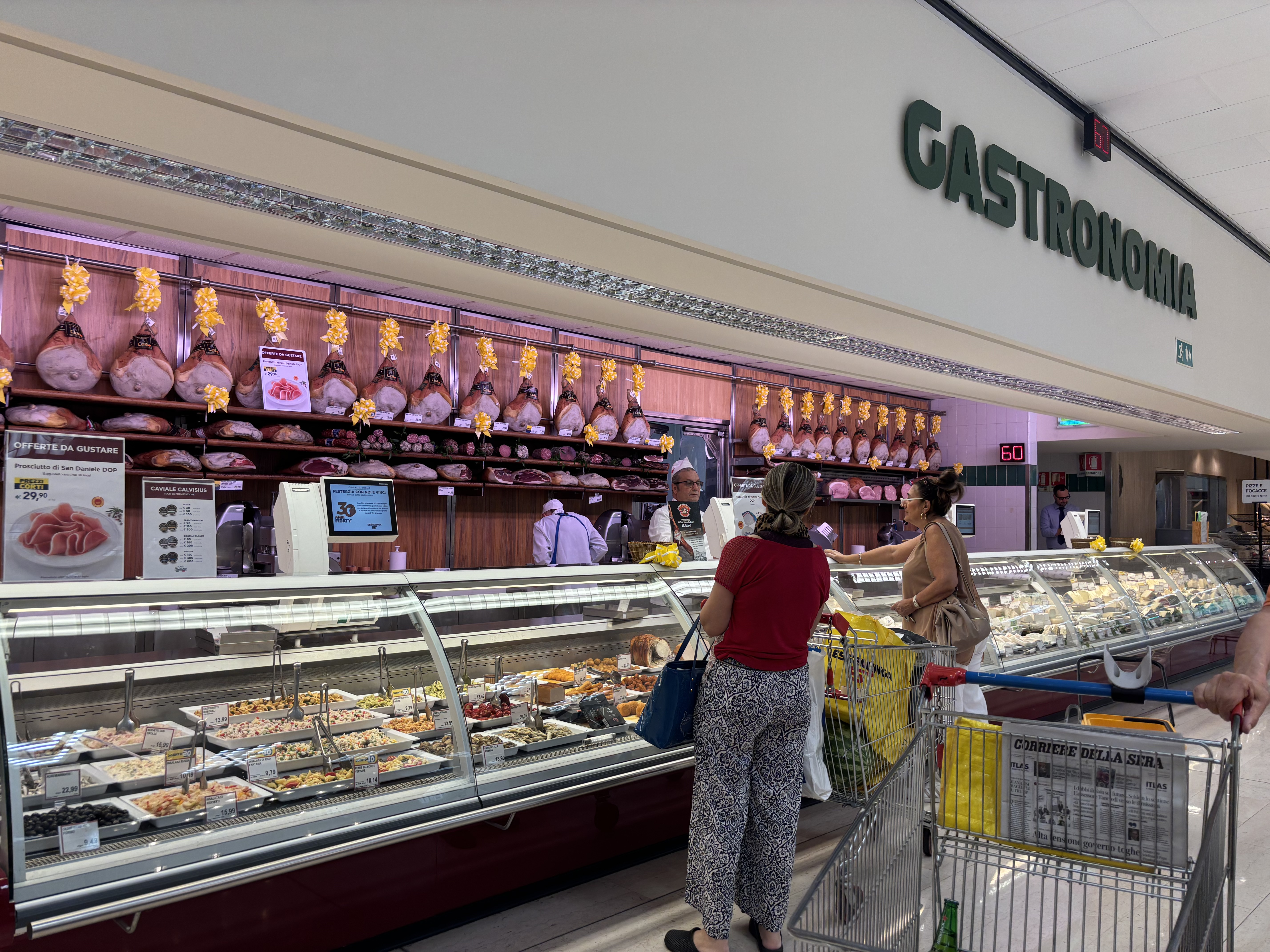 A grocery store deli section labeled "Gastronomia" with displayed meats, a customer at the counter, and an employee serving.