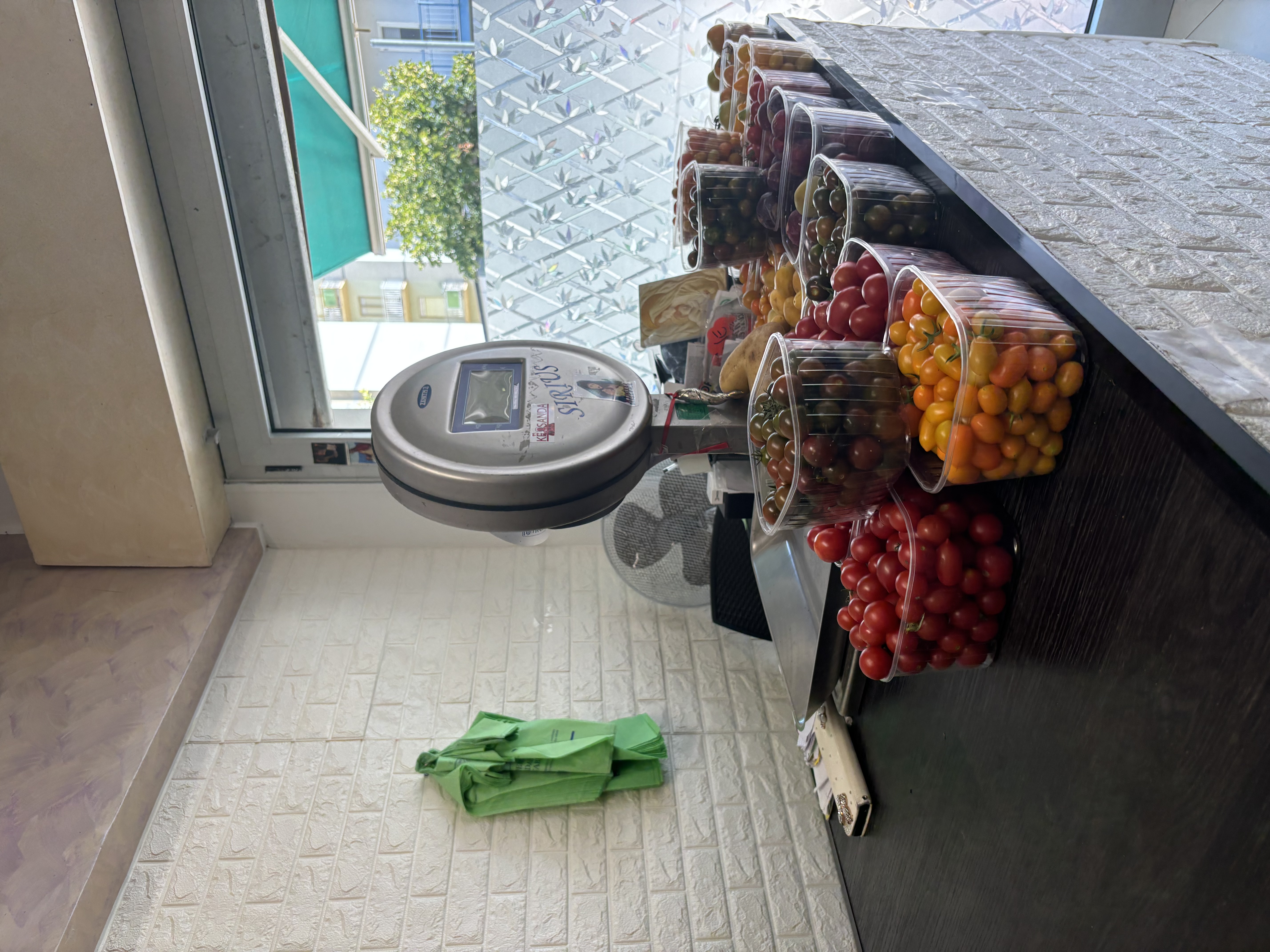 A counter displaying various containers of colorful tomatoes, with a scale and a view of the outdoors through a window.