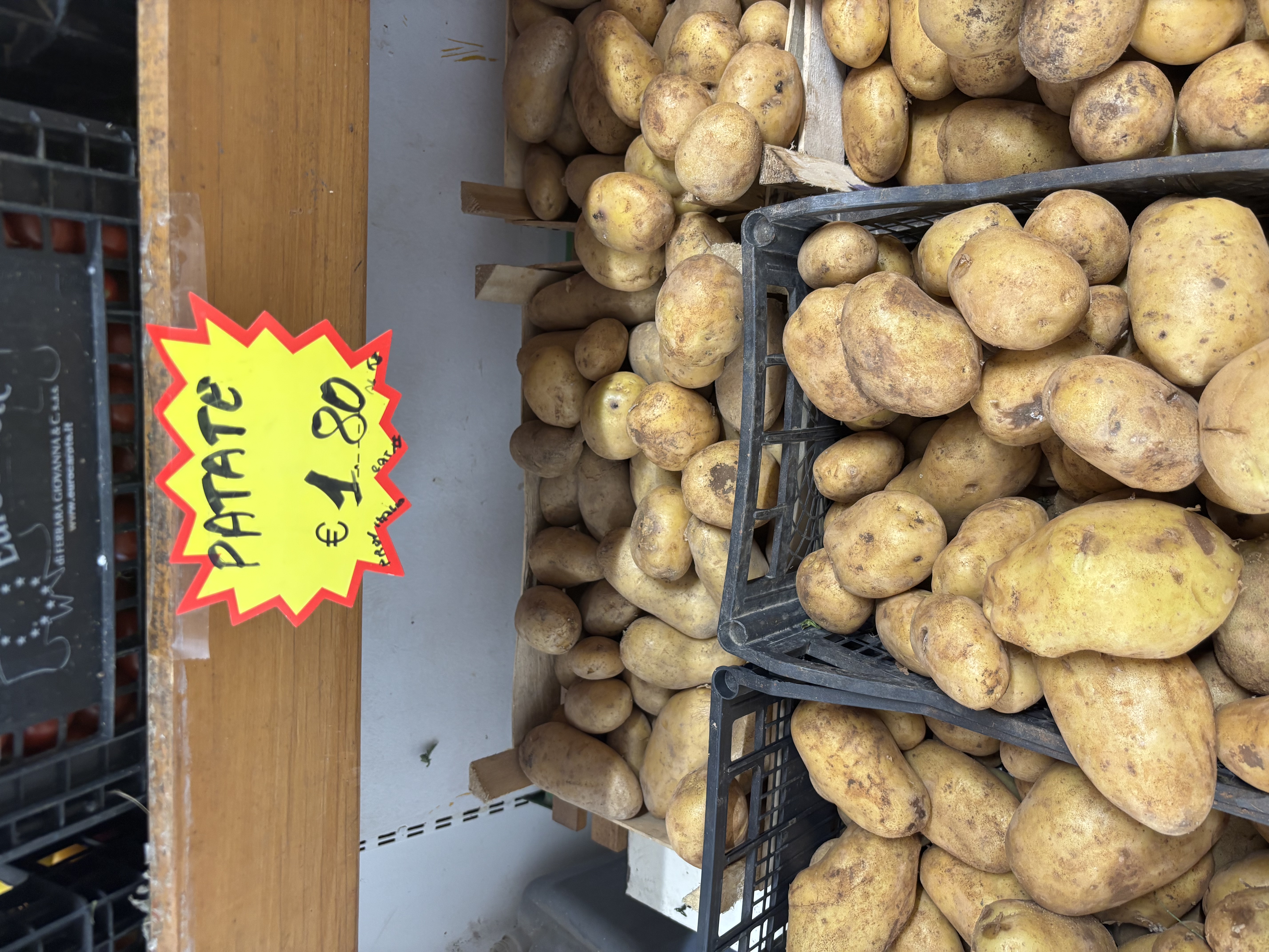 A display of fresh potatoes in crates, with a bright sign reading "PATATE &euro; 1.80."