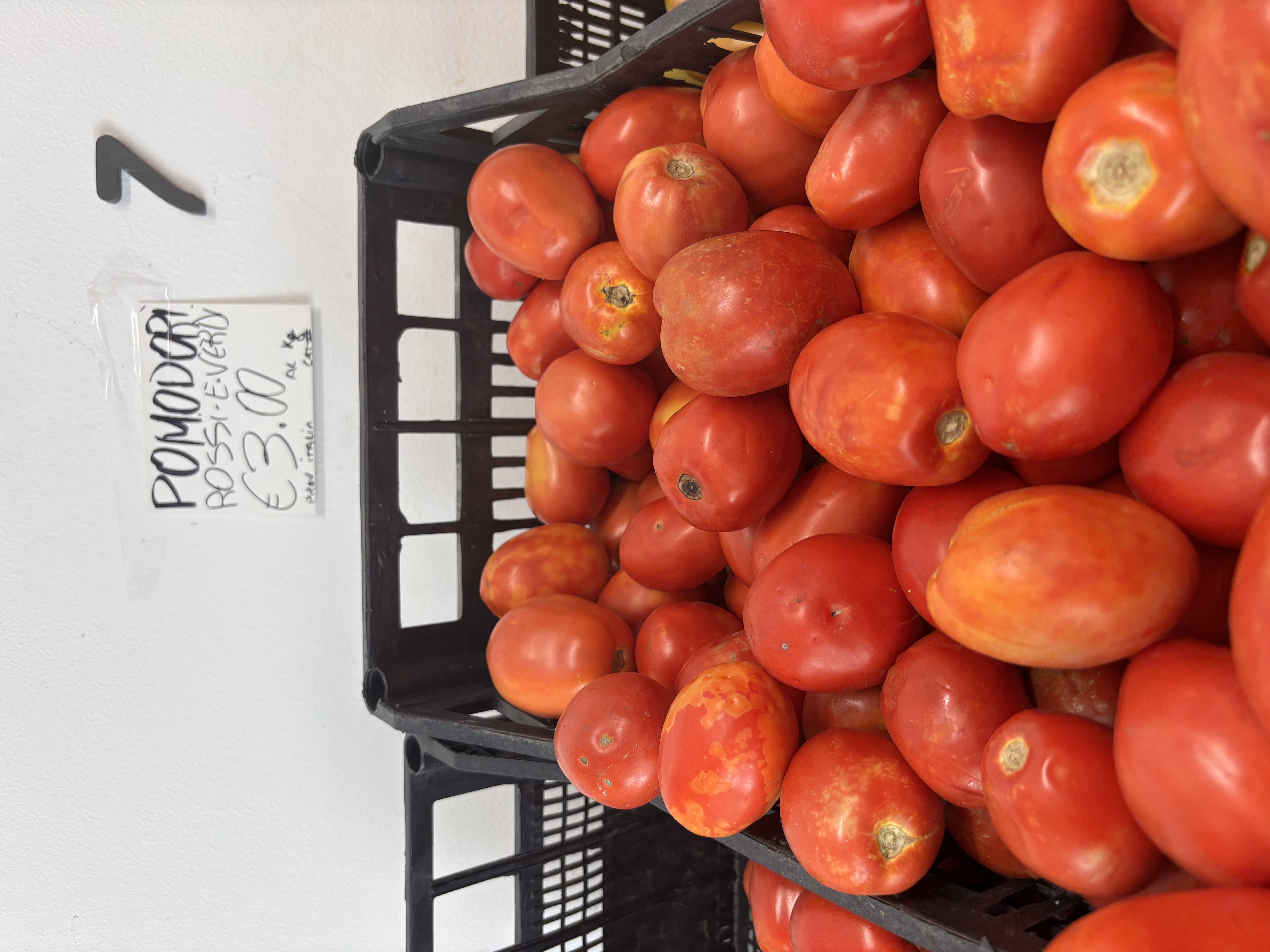 A crate filled with ripe red tomatoes, with a nearby sign reading "Pomodori Rossi - &euro;3.00".