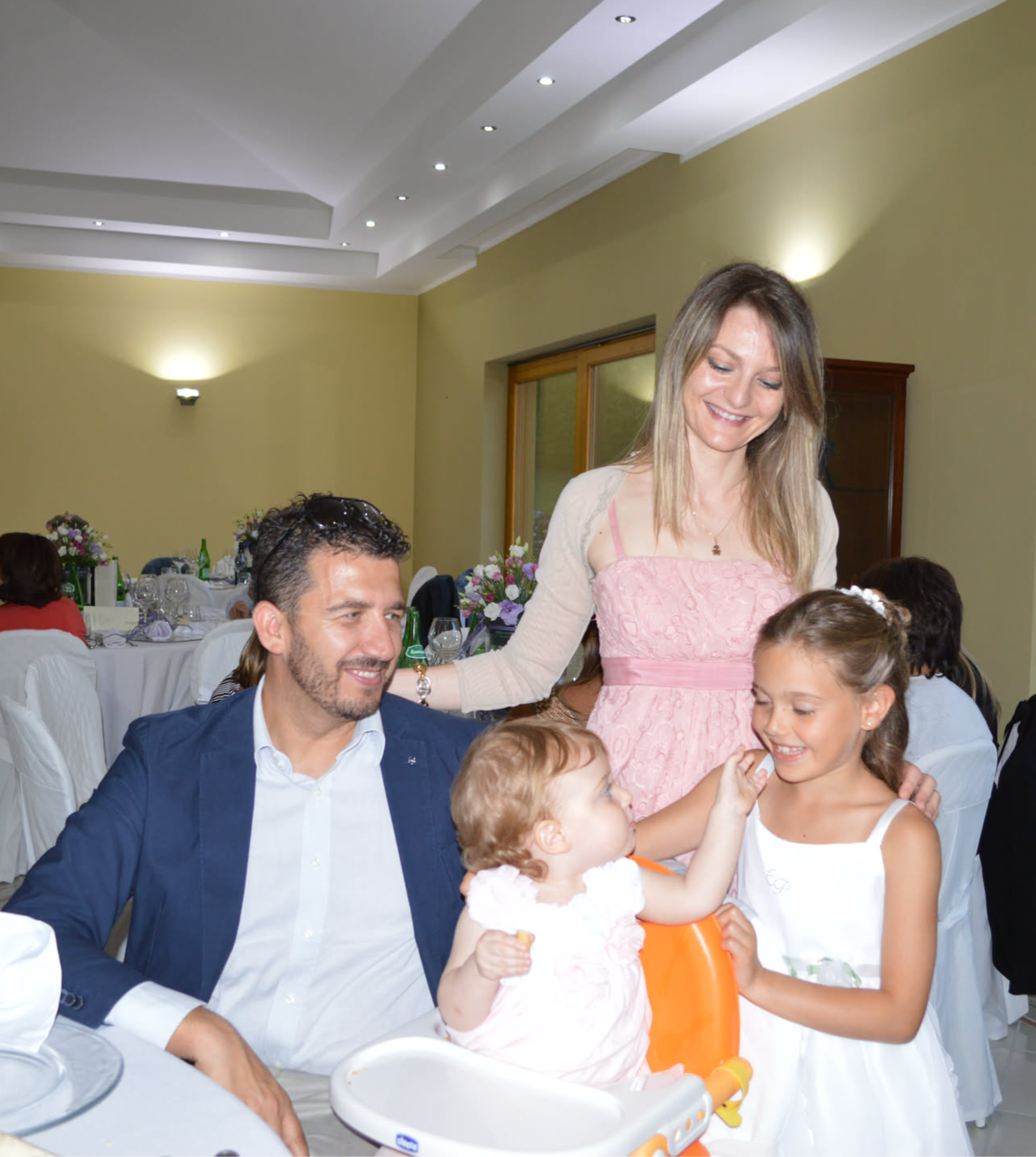A man sits at a table with a baby girl in a high chair, while a woman and a smiling young girl interact beside him.