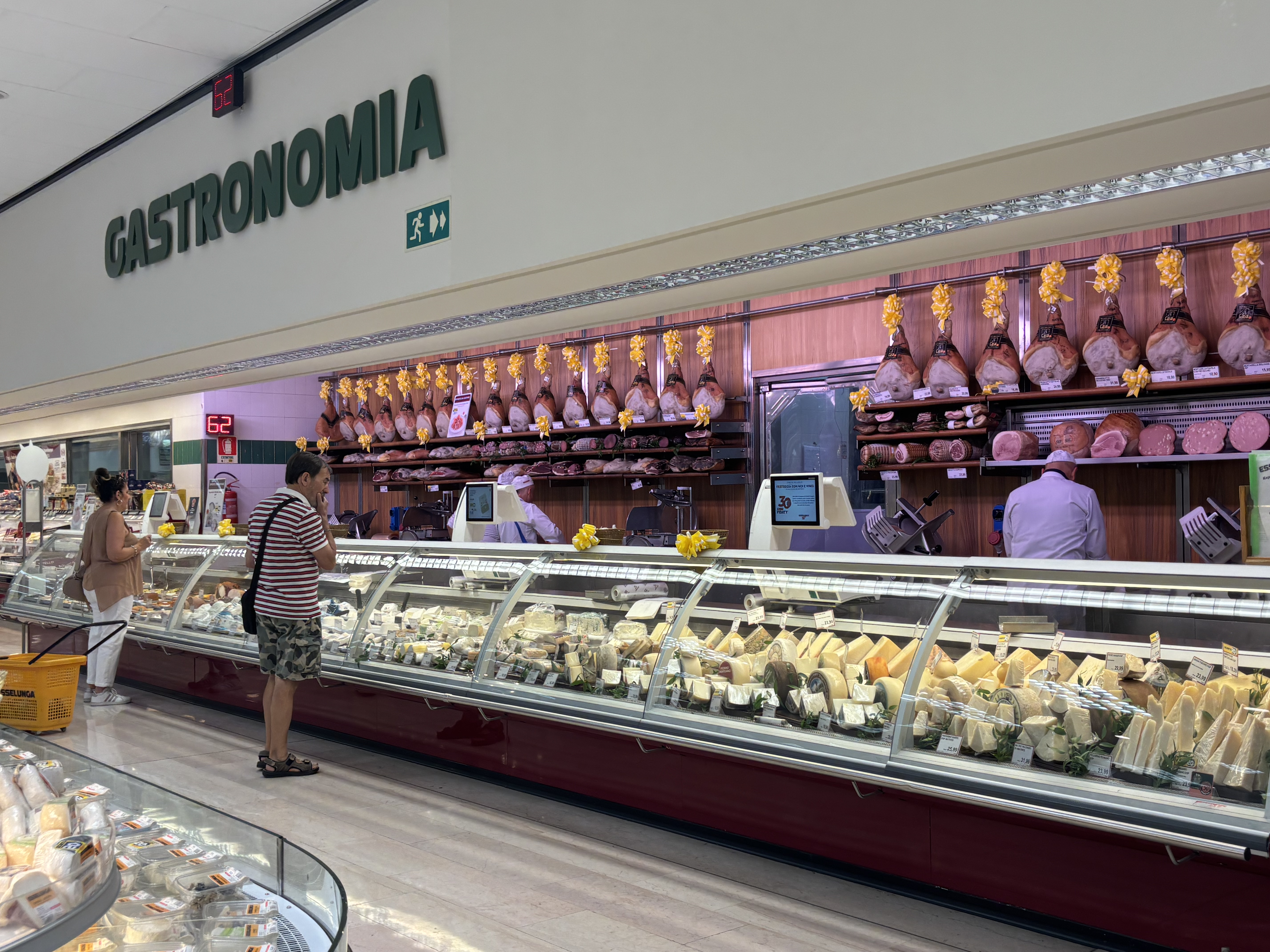 A deli section labeled "Gastronom&iacute;a" displays various meats and cheeses, with customers browsing and staff serving.