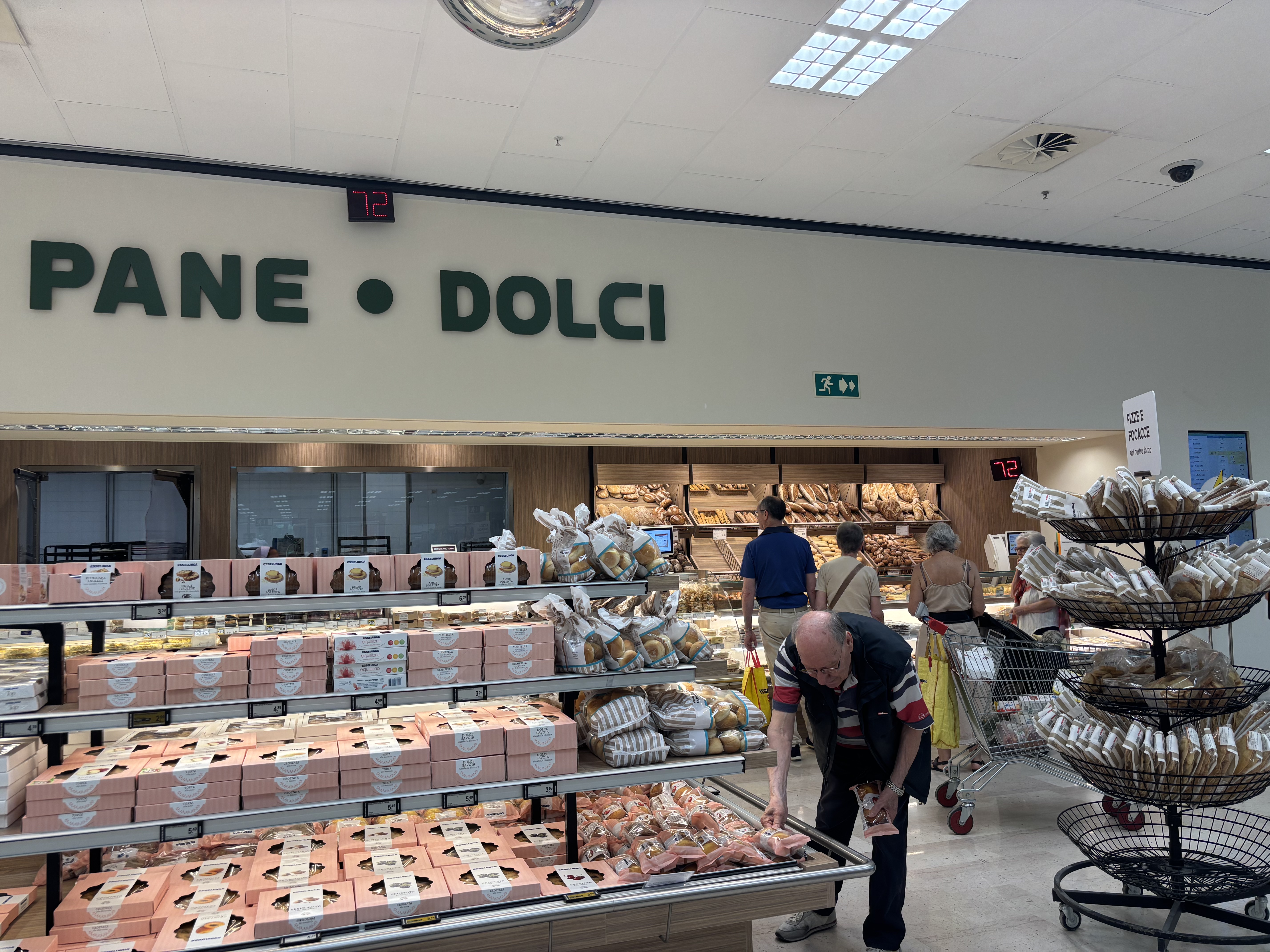 A bakery section in a grocery store, with shelves of baked goods, a sign reading "PANE • DOLCI," and customers browsing.