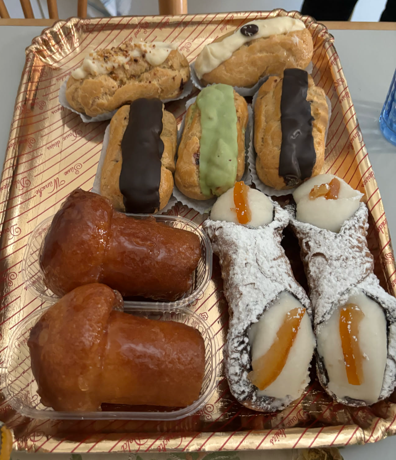 A tray of assorted pastries, including eclairs topped with chocolate, green tea, and cream, plus cannoli and glazed treats.