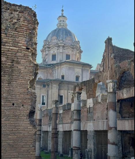 Ruins with stone columns in the foreground, featuring a dome building in the background under a clear blue sky.