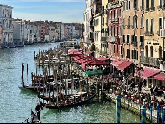 Canal view in Venice with gondolas, colorful buildings, and outdoor dining under red awnings on a sunny day.