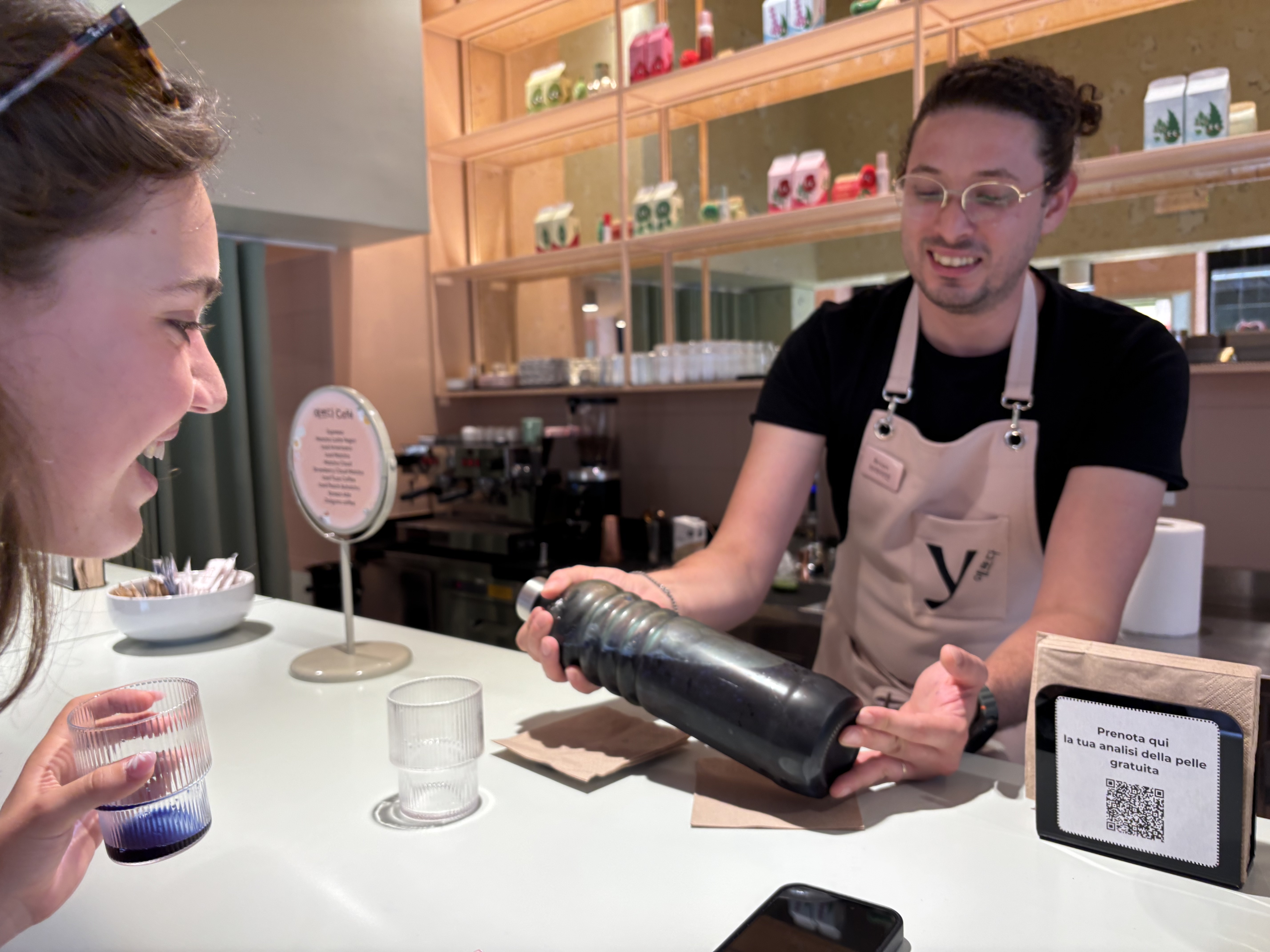 A barista shows a drink in a unique black bottle to a smiling customer at a caf&eacute; counter.