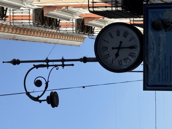 A decorative street clock hangs on a pole against a blue sky, with buildings visible in the background.