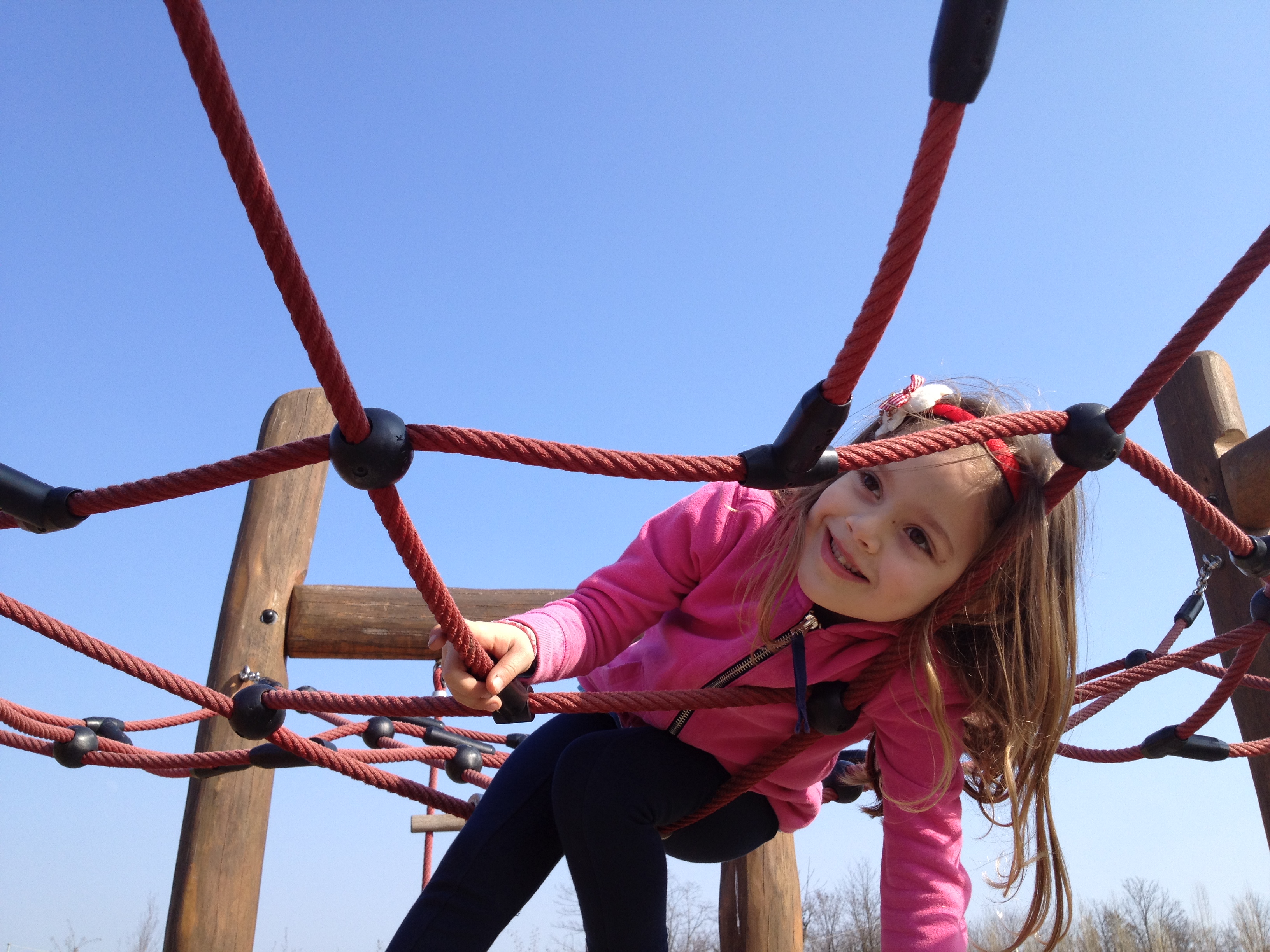 Child smiling while climbing on a red rope structure at a playground against a clear blue sky.