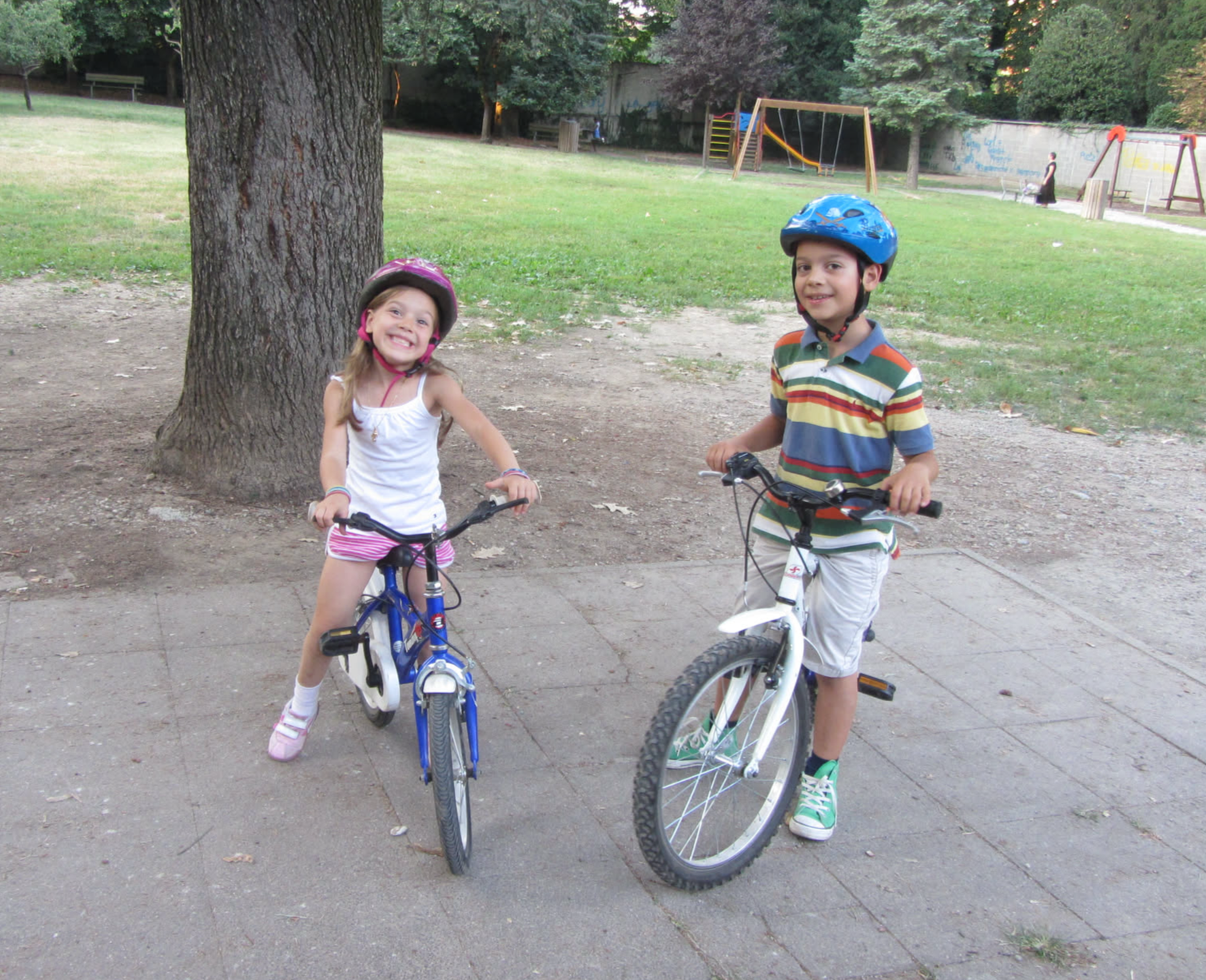 Two children are smiling on bicycles in a park; a girl with a pink helmet and a boy in a striped shirt.