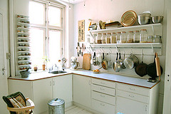 Photo of a white kitchen lit with windows. Rows of glass jars line shelves over the countertop, and a hanging rack of pans and pots appears beneath that.