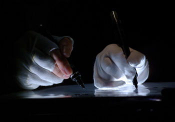 Photograph of two hands writing next to each other, holding black pens and wearing white gloves