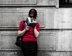 Photo of woman in a red shirt reading a novel while leaning against a gray concrete wall