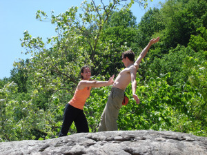 Photo of a woman playfully pretending to shove a shirtless man off of a rock cliff, surrounded by trees