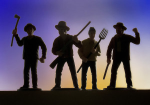 Close-up photo of four plastic figurines, in the shape of men holding axes, pitchforks, and muskets. Figures are in silhouette against a sunset backdrop