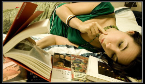 Foto de una mujer acostada de costado leyendo un libro, con un montón de otros libros cerca de su cabeza.