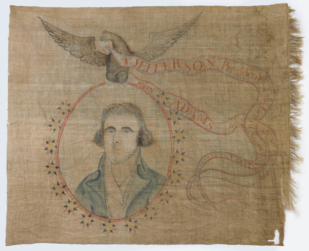 This stitched victory banner celebrating the presidential election of Thomas Jefferson depicts a portrait of Jefferson in the center of a red circle that is outlined by yellow and blue stars. An eagle sits atop the circle with a ribbon in its mouth that reads, &ldquo;T. Jefferson, President of the United States of America. John Adams is no more.&rdquo;