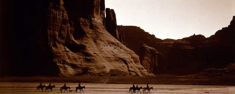 Seven silhouetted riders move through a canyon with tall cliffs behind them.