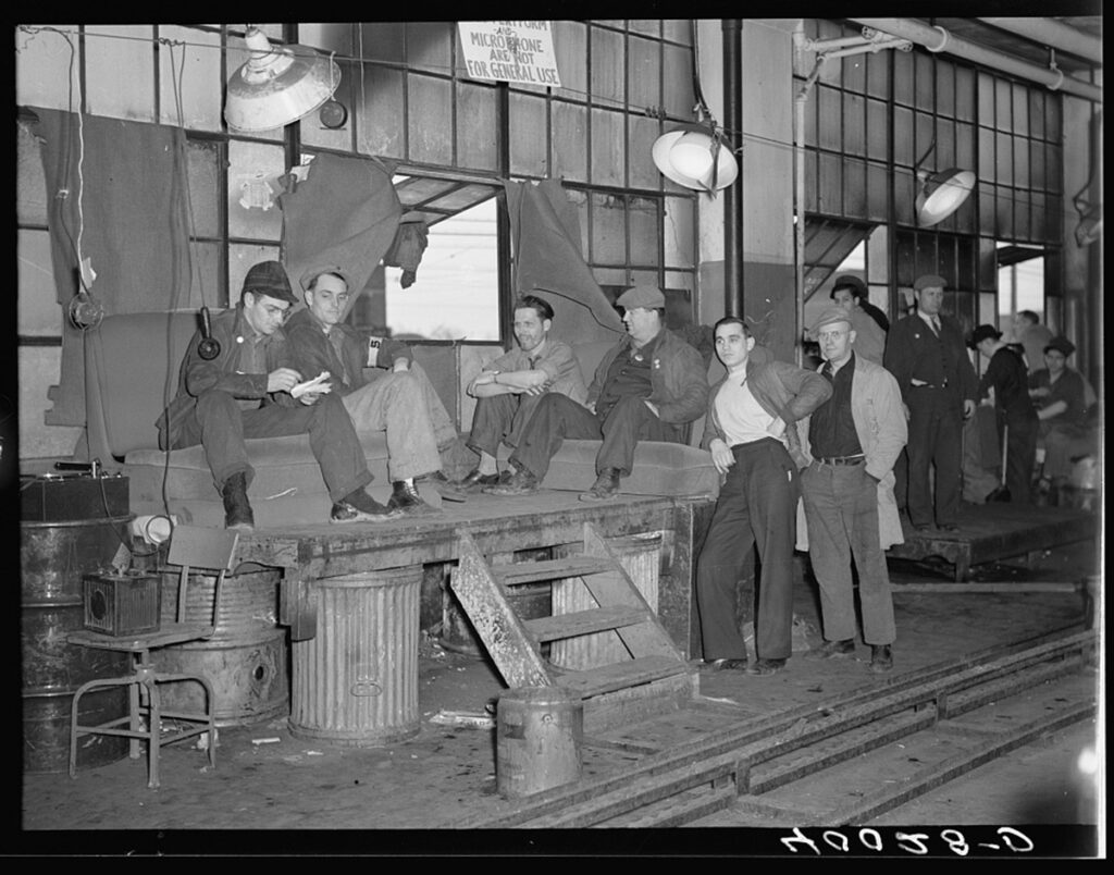 Men lounge outside of a factory building. 