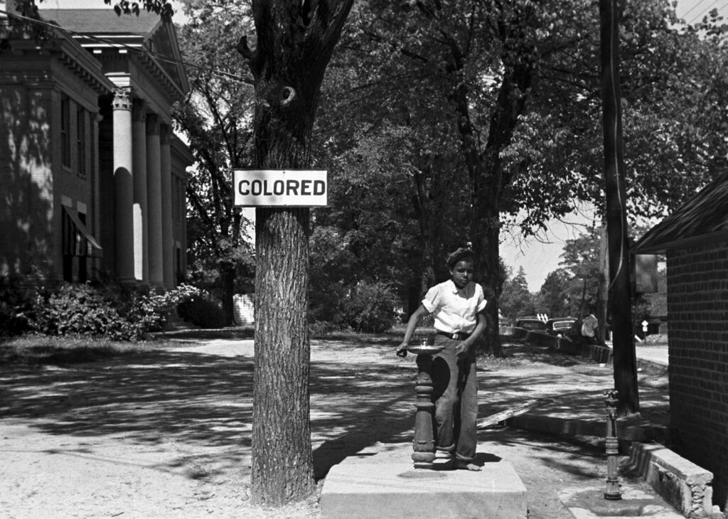 A boy drinks from an outdoor water fountain in front of a tree with a sign reading &ldquo;Colored.&rdquo;