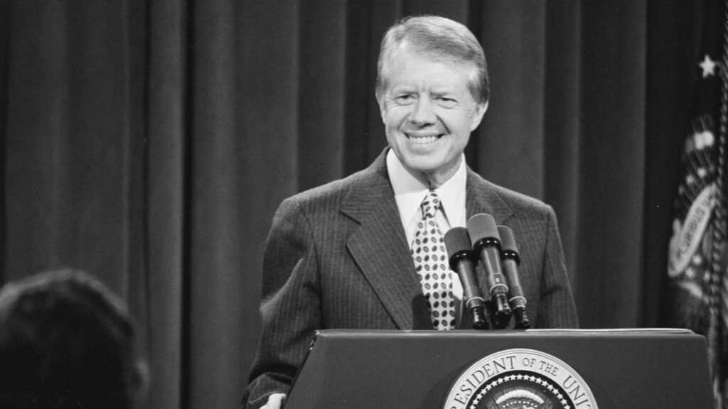 A man in a suit stands before a lectern with microphones.
