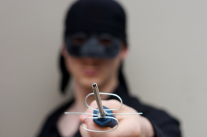 Photo foregrounding a homemade sword held by a man dressed in all black, wearing a mask