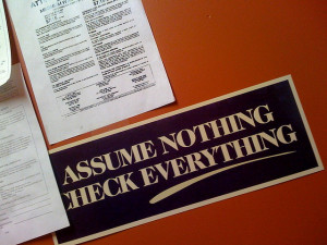 Photo of papers on a desk, with a bumper sticker reading "Assume Nothing Check Everything"