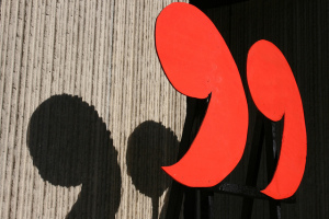 Photo of quotation mark sculpture. The punctuation marks are bright red, standing upright on thin rods. They cast dark shadows on the stone wall behind them