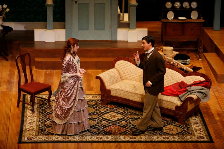 una sala de estar de la época victoriana con una mujer vestida rosa (Nora) que parece ser conferenciada por un hombre de traje (su esposo, Torvald)