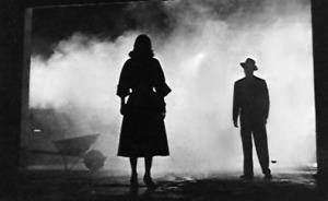 A still shot from a black and white 1940's movie is shown. We see the profile of a woman's full body in the center of the screen, and a man further away on the right. Mist billows behind them, and the contrast is high between black and white.