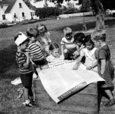 Black and white photo of several children clustered around a poster of the Declaration of Human Rights on a milk crate in a backyard play area