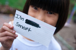 Girl holding an index card in front of her face, reading "I'm [scribbled out word] smart"