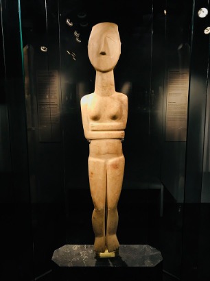 A statue in the Cycladic style, a human form with arms wrapped around its center and lacking detail.