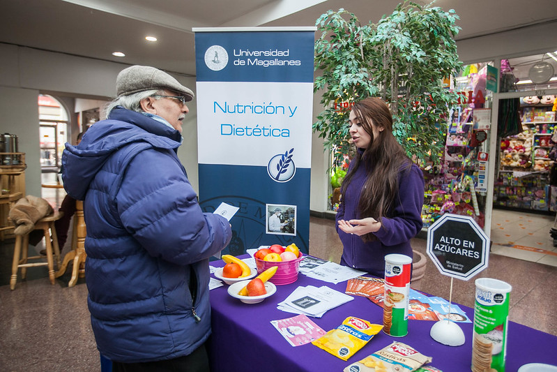 A woman stands behind a table on which pamphlets and food samples are laid out. She is talking to an older man. The sign behind her says "Nutrición y Dietética."