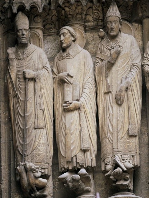 A sculpture of three saints. The saints are a part of the wall they are carved from. None of their arms extend out from their forms; the sculptures are all very solid.