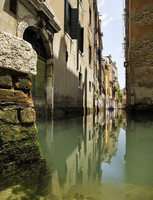 A building can be seen nearly perfectly reflected in the canal; however, the water distorts and discolors the image.